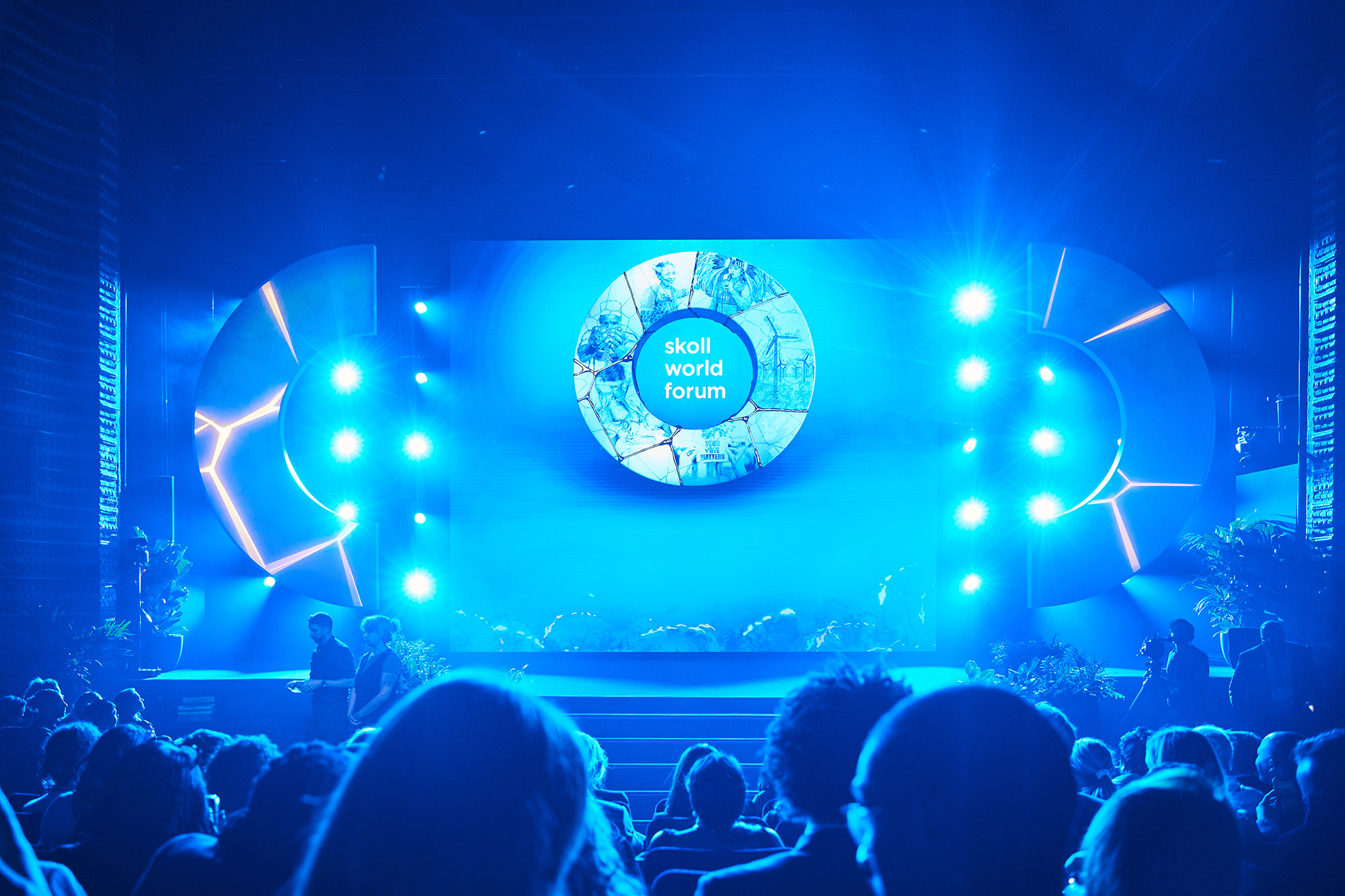 The Skoll World Forum 2025 stage lit in blue, with the kintsugi ring on the screen between two gold-veined stage pieces and the audience in the foreground
