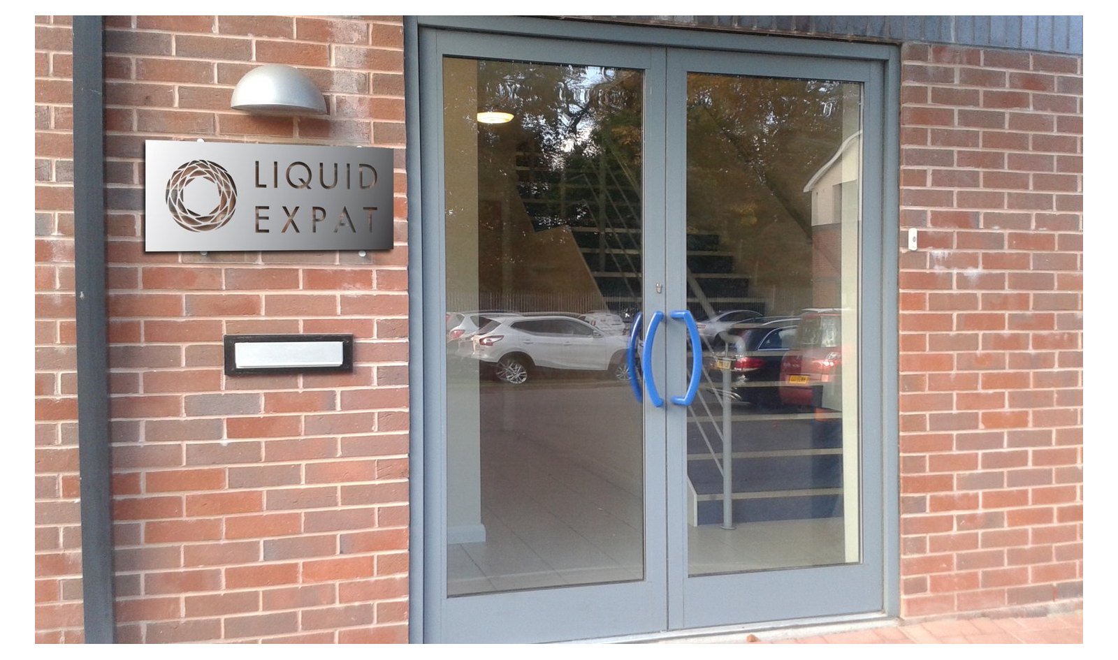The Manchester office entrance with the Liquid Expat mark cut through a brushed stainless plate mounted on a red brick wall beside glass doors.