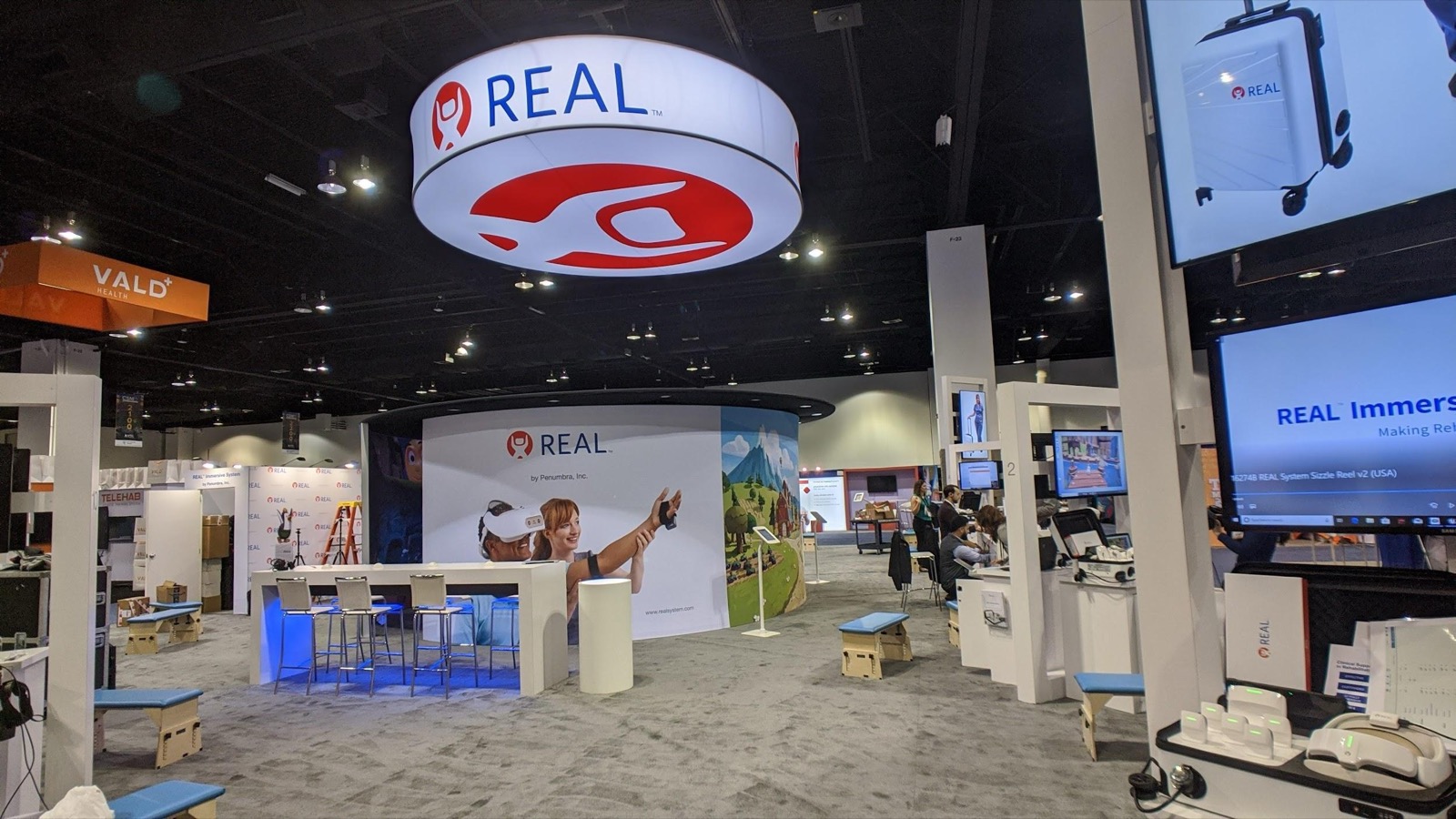 The REAL System stand at the APTA conference: a circular overhead sign, demo stations with headsets, and screens running the product reel.
