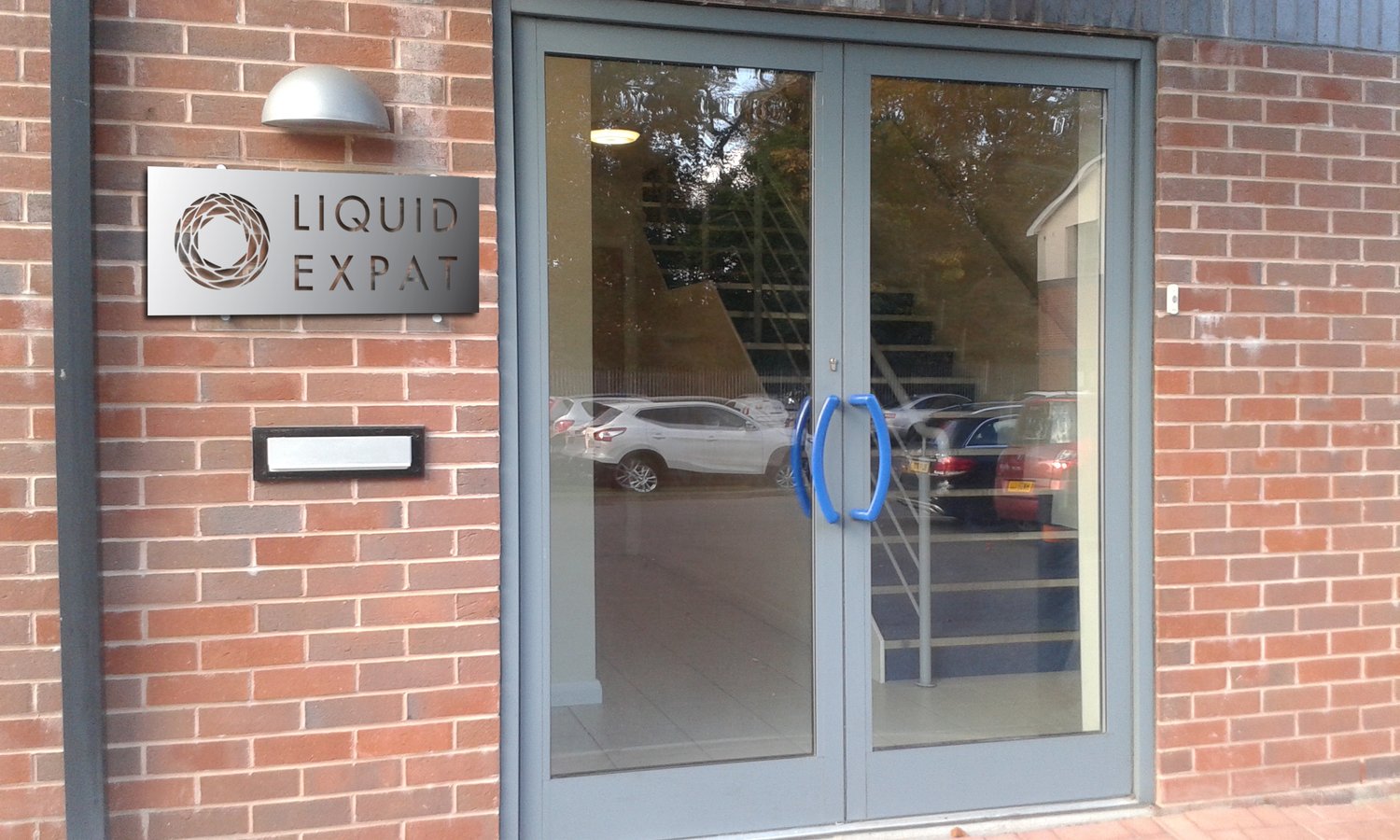 The Manchester office entrance: a brushed metal plate mounted on a red brick wall beside glass doors, with the Liquid Expat logomark and wordmark cut through the metal.